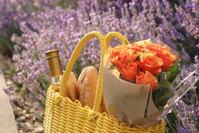 Yellow wicker bag with beautiful roses, bottle of wine and baguettes near lavender flowers outdoors, closeup