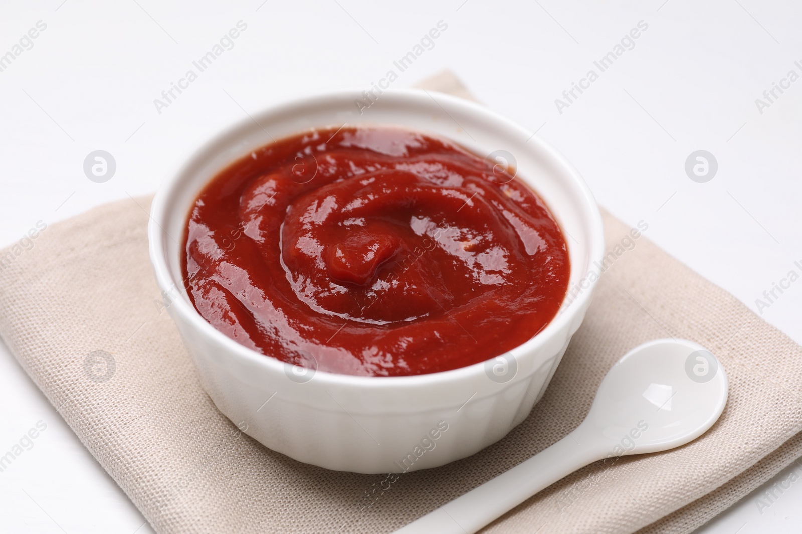 Photo of Organic ketchup in bowl and spoon on white table, closeup. Tomato sauce