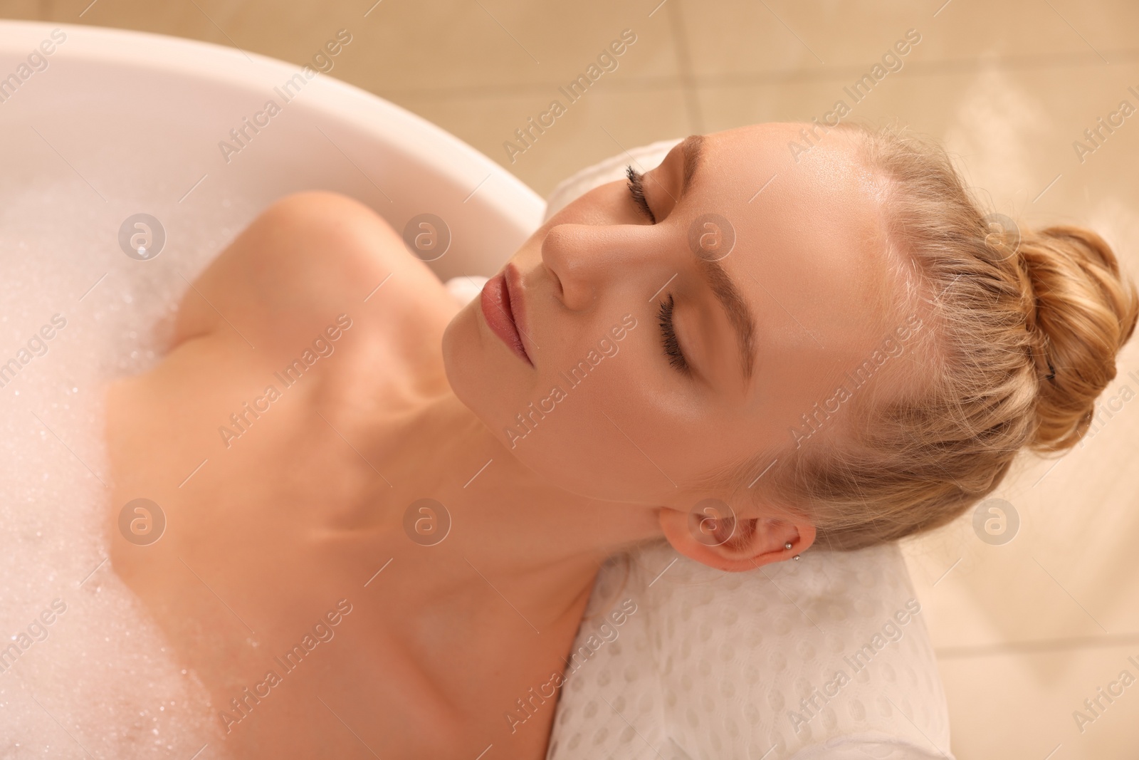 Photo of Young woman using pillow while enjoying bubble bath indoors