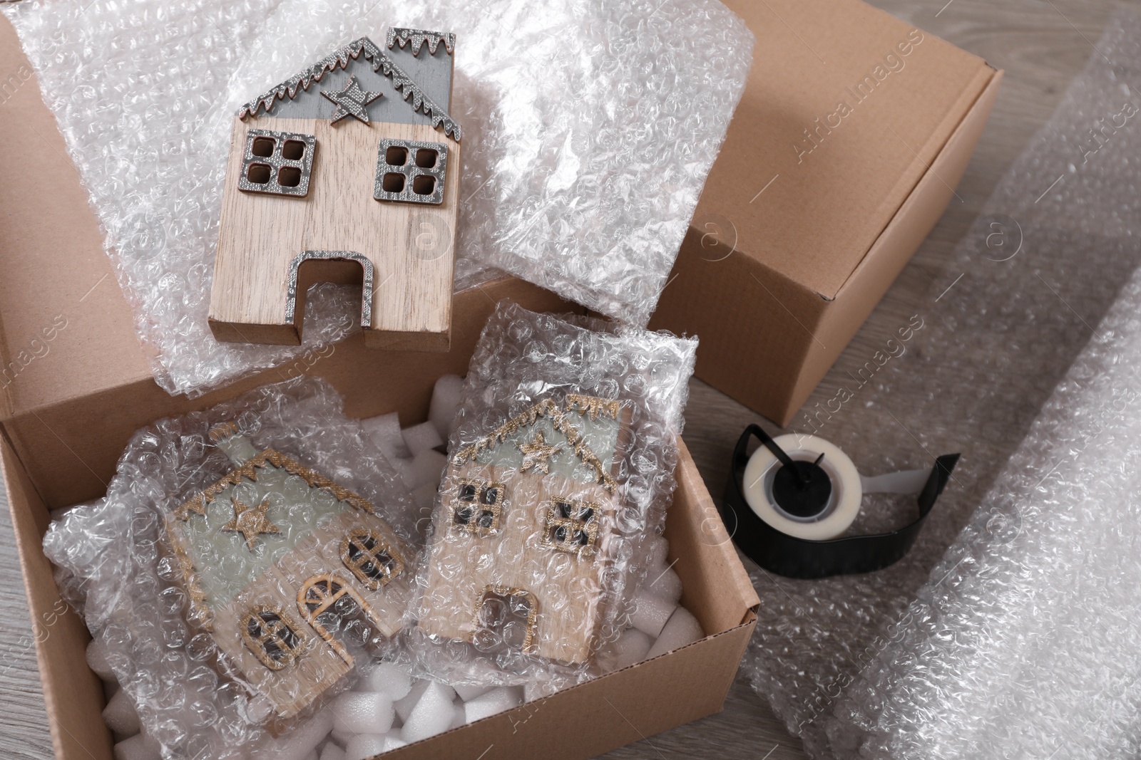 Photo of Open box with decorative house figures, adhesive tape, foam peanuts and bubble wrap on wooden table