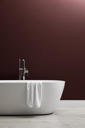 Modern ceramic bathtub with towel near burgundy wall indoors