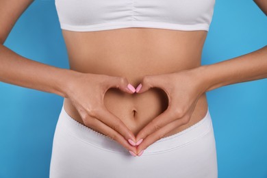 Woman in underwear making heart with hands on her belly against light blue background, closeup. Healthy stomach