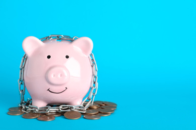 Photo of Piggy bank  with steel chain and coins on light blue background, space for text. Money safety concept