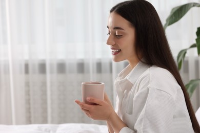 Happy woman with cup of drink on bed at home, space for text. Lazy morning