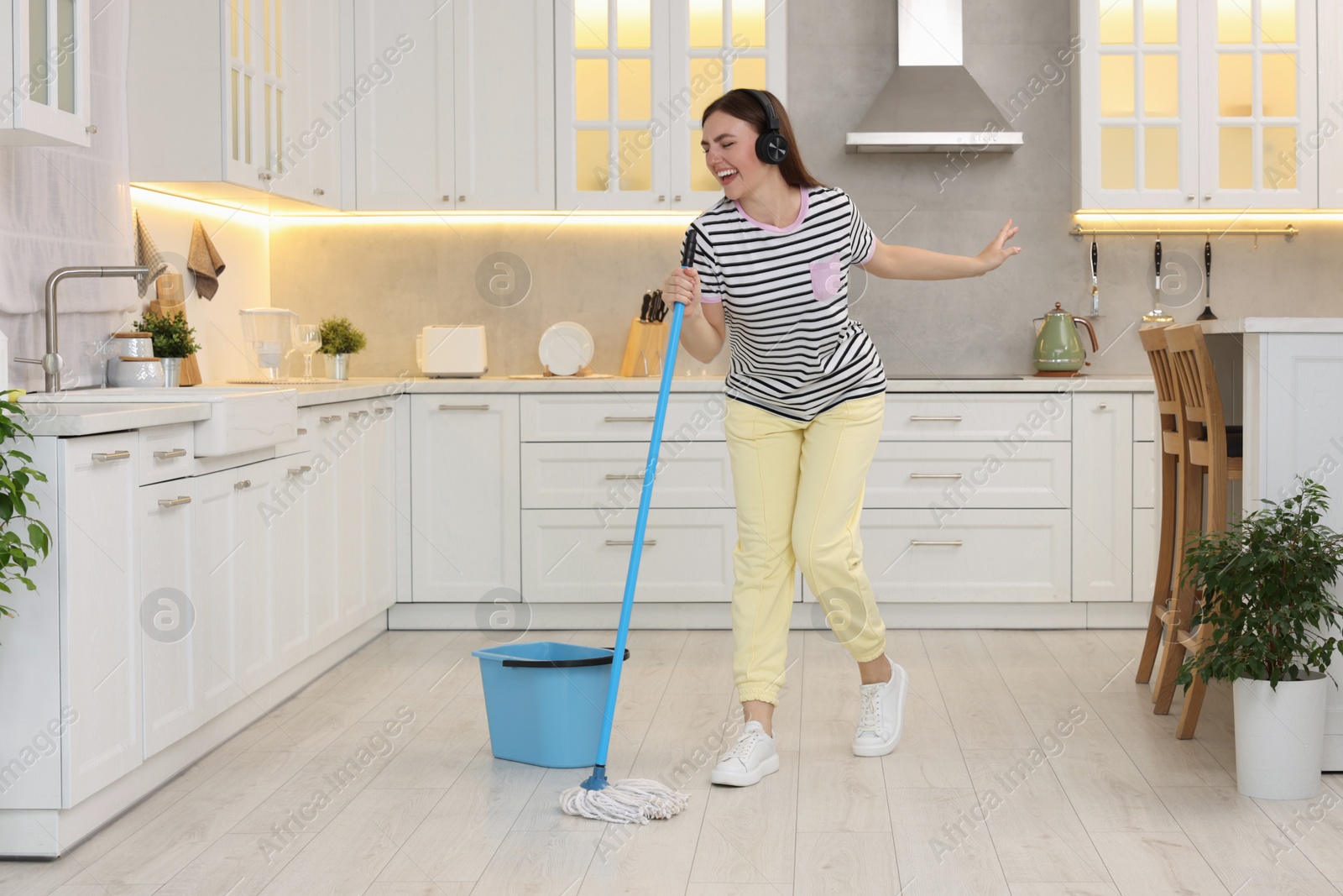 Photo of Enjoying cleaning. Happy woman in headphones dancing with mop in kitchen