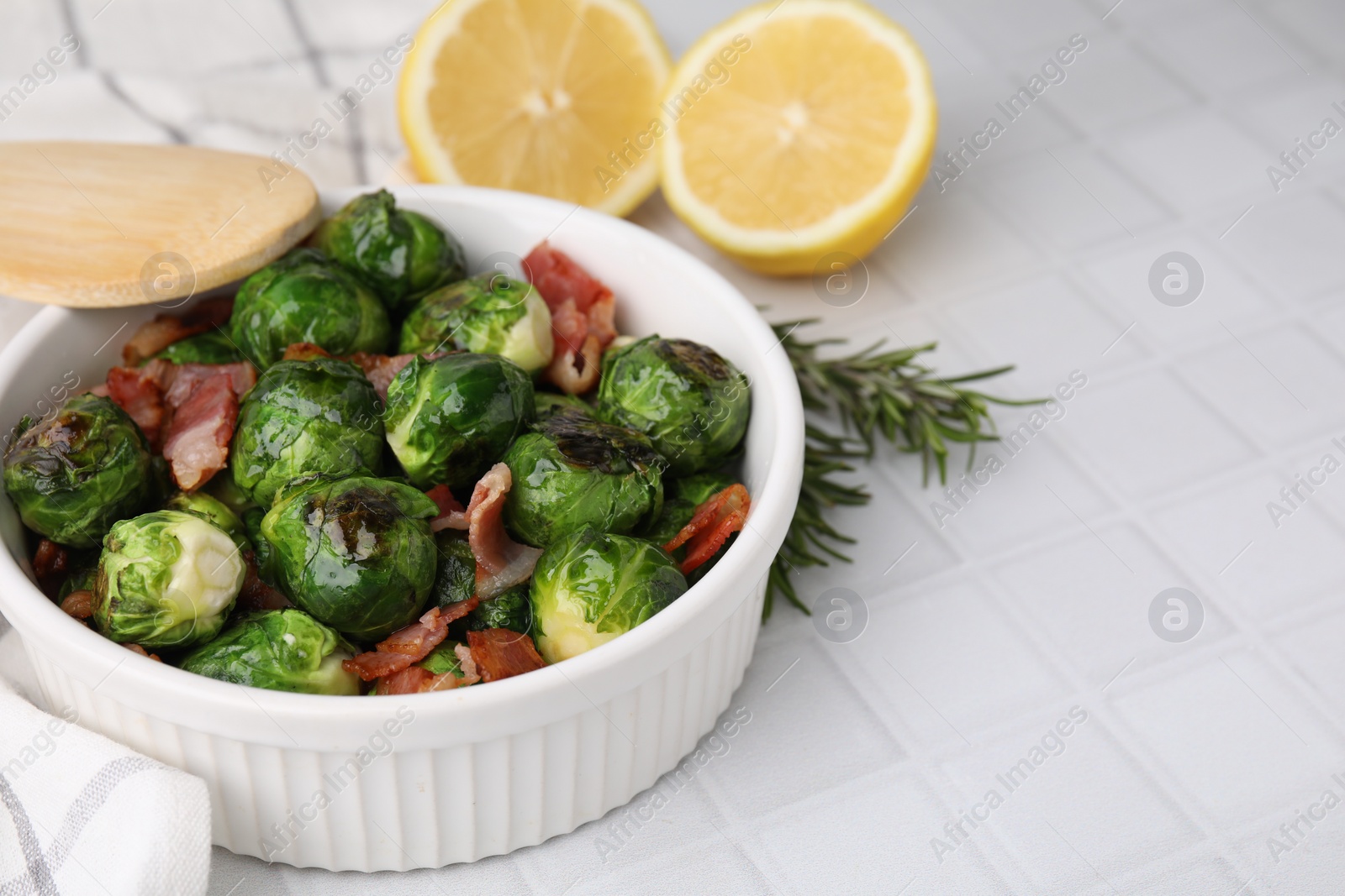 Photo of Delicious roasted Brussels sprouts, bacon, lemon and rosemary on white tiled table, closeup. Space for text