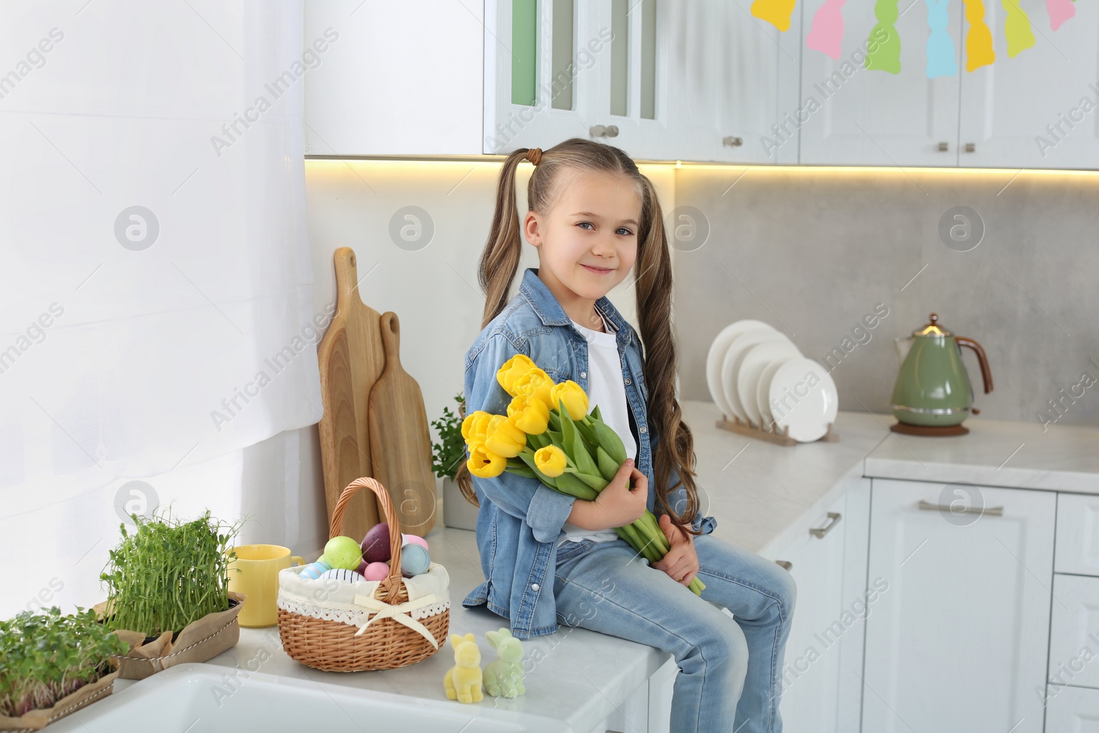 Photo of Cute girl with yellow tulips and wicker basket full of Easter eggs in kitchen