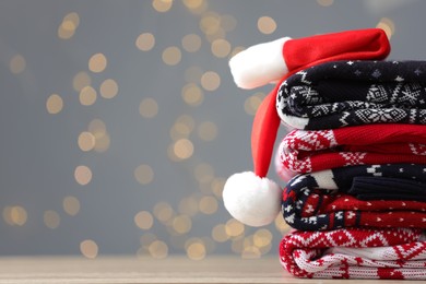 Photo of Stack of different Christmas sweaters and Santa hat on table against grey background with blurred lights. Space for text