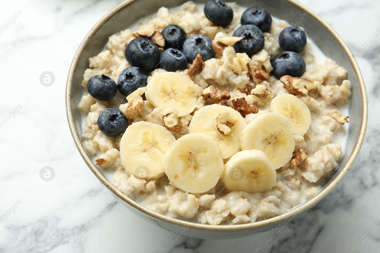 Photo of Tasty oatmeal with banana, blueberries, walnuts and milk served in bowl on white marble table, closeup