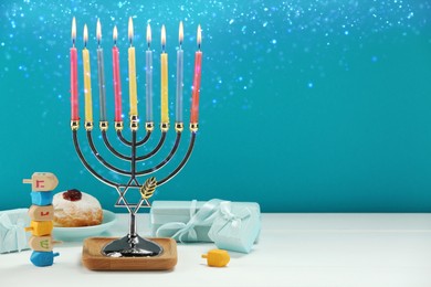 Image of Hanukkah celebration. Menorah with burning candles, dreidels, gift boxes and donut on white wooden table against light blue background, space for text