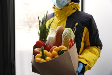 Photo of Courier in medical mask holding paper bag with groceries at doorway, closeup. Delivery service during quarantine due to Covid-19 outbreak