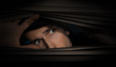 Image of Worried man looking through window blinds into darkness. Paranoia concept