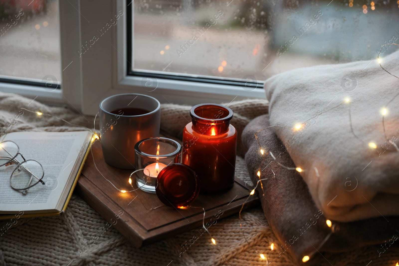 Photo of Cup of hot drink, open book and Christmas lights on knitted blanket near window. Cozy season