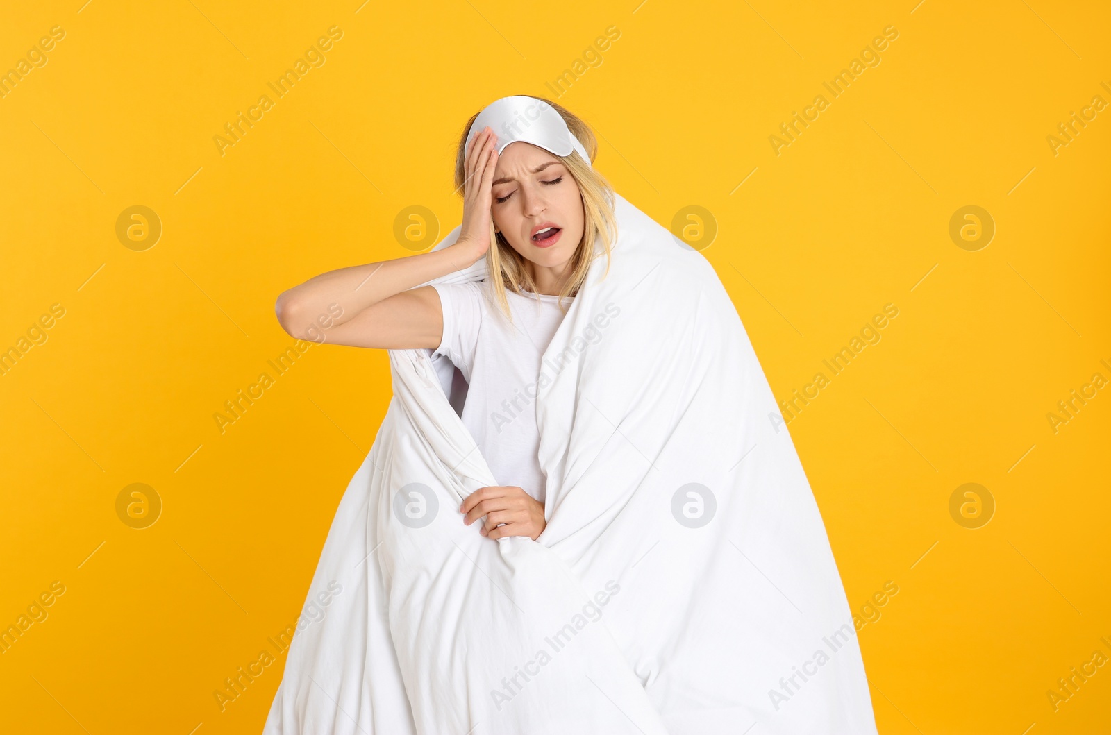 Photo of Young woman in sleeping mask wrapped with soft blanket yawning on yellow background