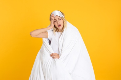 Photo of Young woman in sleeping mask wrapped with soft blanket yawning on yellow background