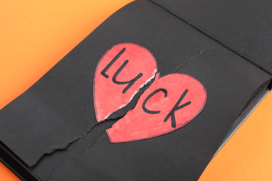Photo of Torn word LUCK with heart written in notebook on orange background, closeup
