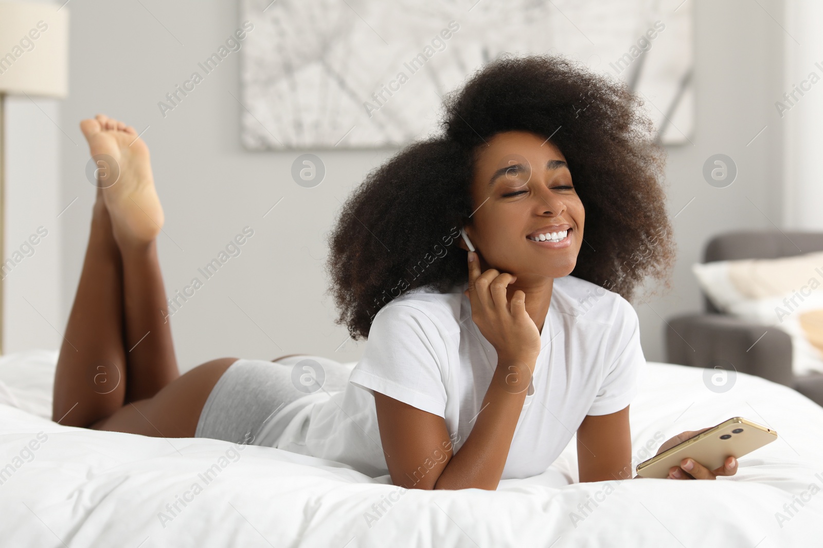 Photo of Beautiful woman in stylish underwear and t-shirt listening to music through wireless earphones on bed indoors