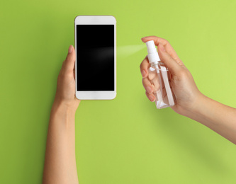 Image of Woman sanitizing smartphone with antiseptic spray on green background, closeup. Be safety during coronavirus outbreak 