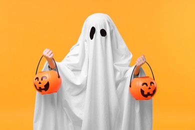 Photo of Child in white ghost costume holding pumpkin buckets on orange background. Halloween celebration
