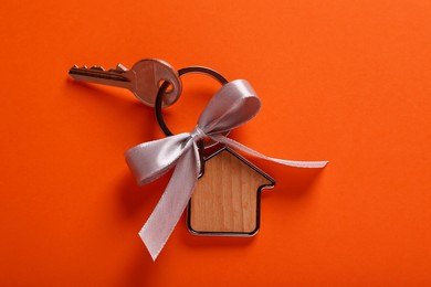 Key with trinket in shape of house and grey bow on orange background, top view. Housewarming party