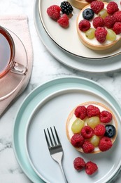 Photo of Delicious tartlets with berries and tea on white marble table, flat lay