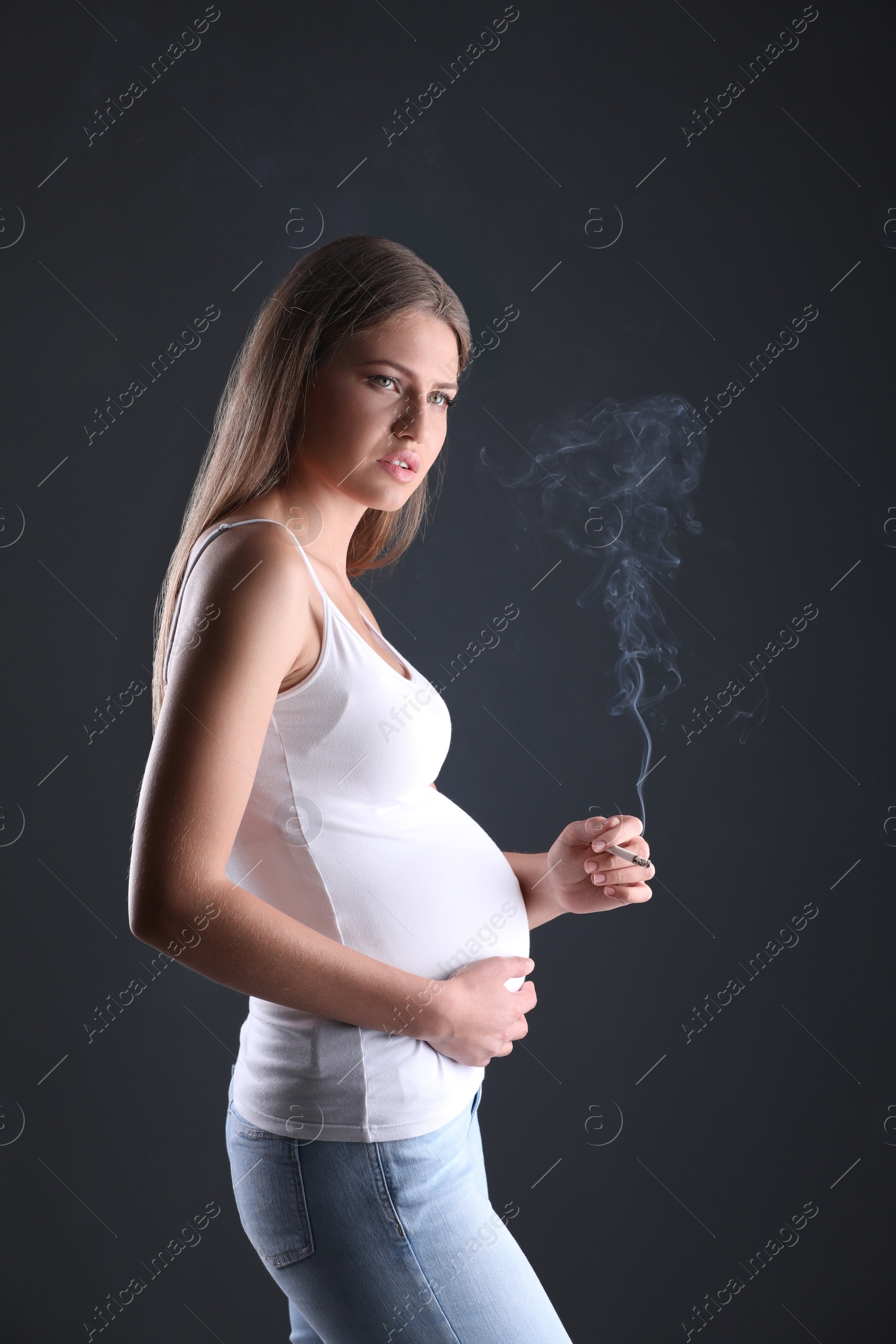 Photo of Young pregnant woman smoking cigarette on dark background. Harm to unborn baby