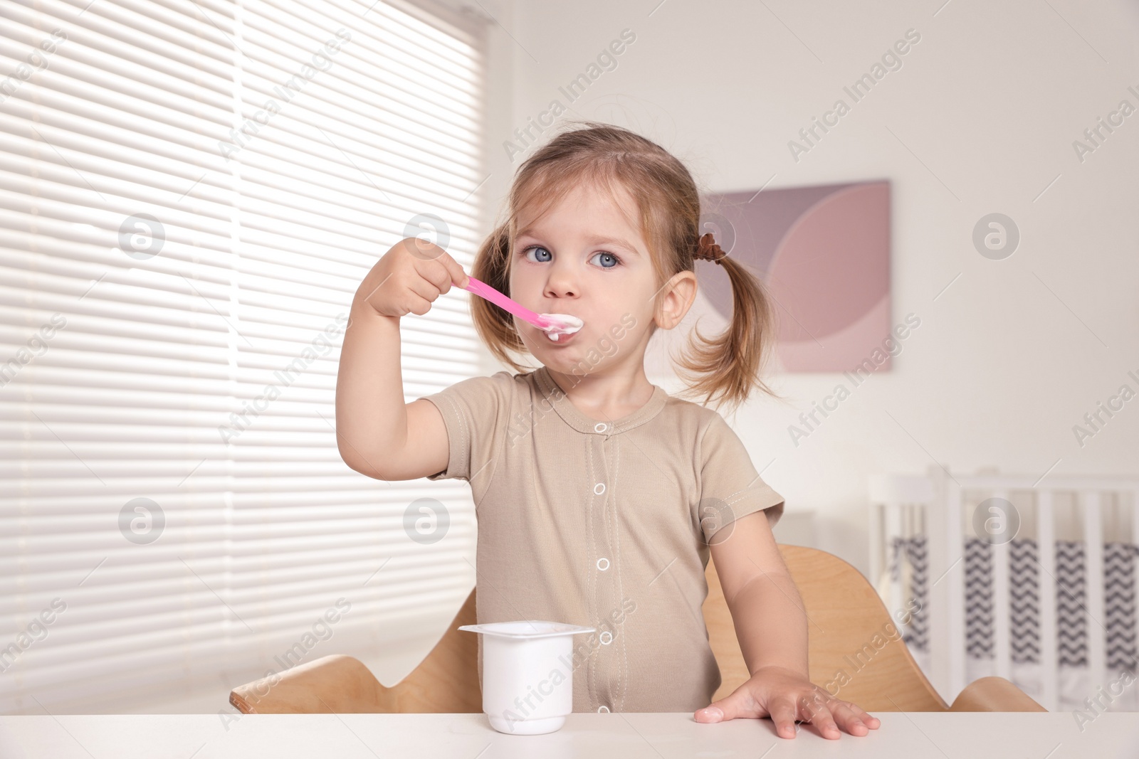 Photo of Cute little child eating tasty yogurt from plastic cup with spoon at white table indoors