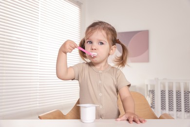 Cute little child eating tasty yogurt from plastic cup with spoon at white table indoors