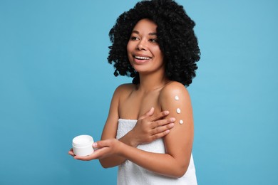 Young woman applying body cream onto shoulder on light blue background