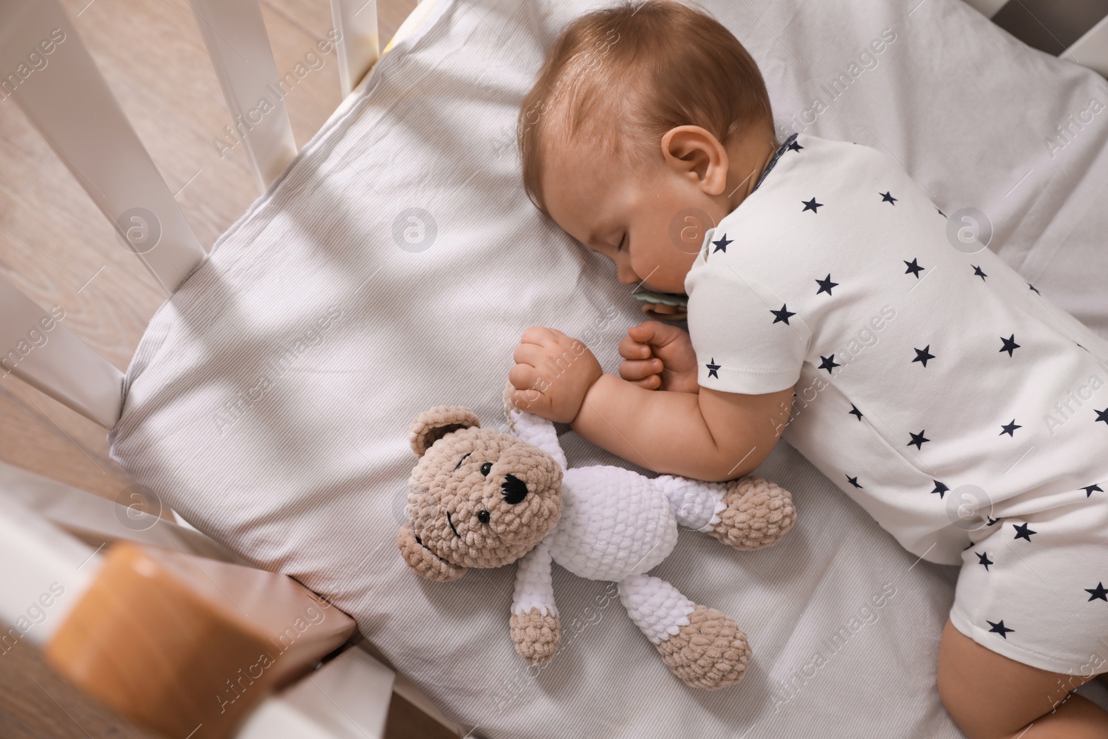 Photo of Adorable little baby with pacifier and toy sleeping in crib indoors, above view