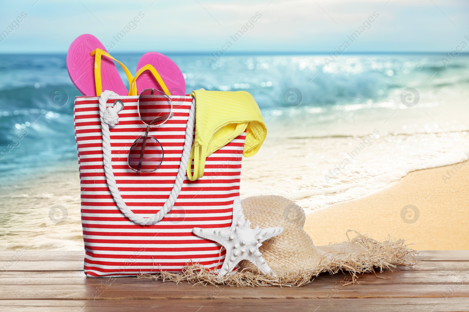 Image of Stylish striped bag and other beach accessories on wooden table near sea