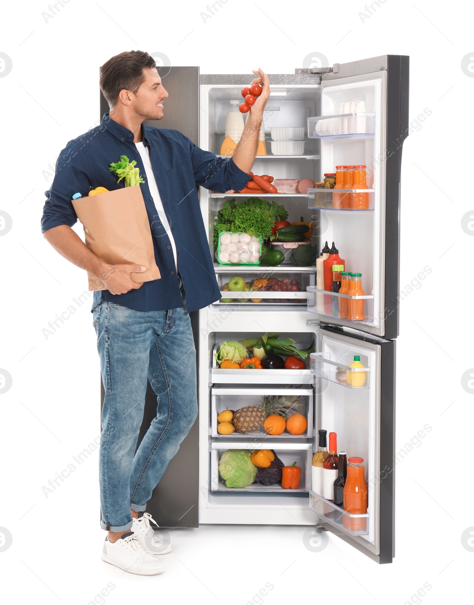 Photo of Man with bag of groceries and tomatoes near open refrigerator on white background