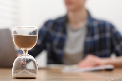 Photo of Hourglass with flowing sand on desk. Man taking notes while using laptop indoors, selective focus