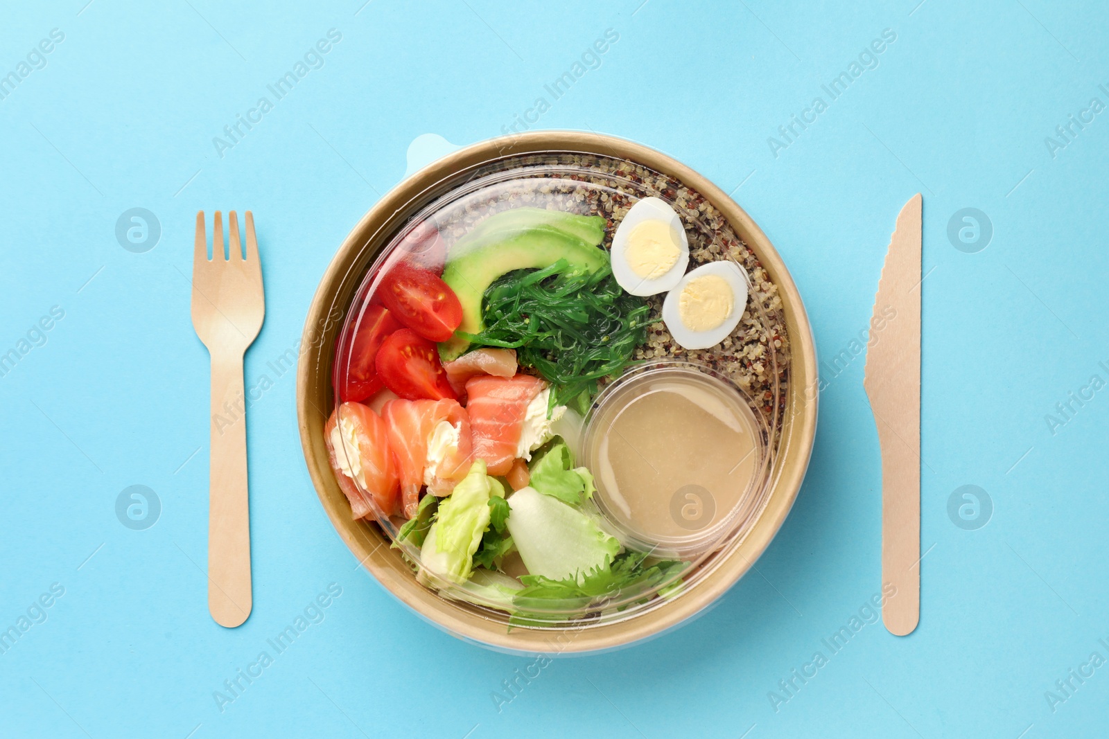 Photo of Tasty food in container with wooden fork and knife on light blue background, flat lay