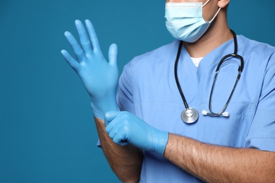 Doctor in protective mask putting on medical gloves against blue background, closeup
