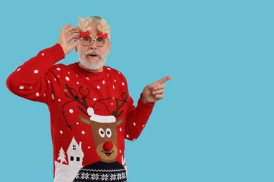Surprised senior man in Christmas sweater and party glasses pointing at something on light blue background. Space for text