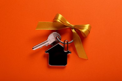 Key with trinket in shape of house and yellow bow on orange background, top view. Housewarming party