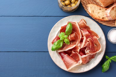Photo of Slices of tasty cured ham, basil, bread and olives on blue wooden table, flat lay. Space for text