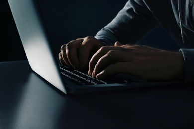 Man using laptop at table on dark background, closeup. Criminal activity
