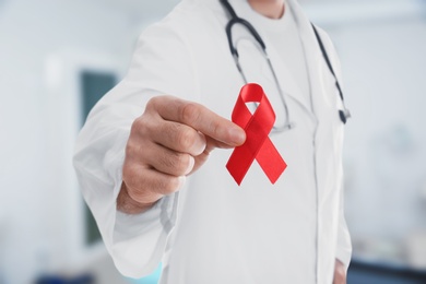 Image of Doctor holding red awareness ribbon on blurred background, closeup. World AIDS disease day