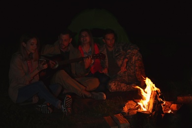 Photo of Young man playing guitar for friends near bonfire at night. Camping season