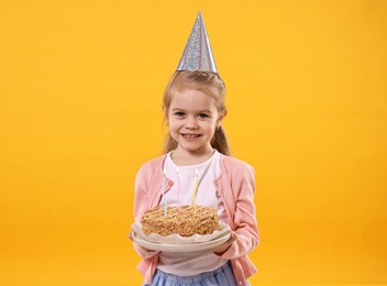 Birthday celebration. Cute little girl in party hat holding tasty cake with burning candles on orange background