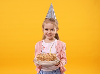 Photo of Birthday celebration. Cute little girl in party hat holding tasty cake with burning candles on orange background