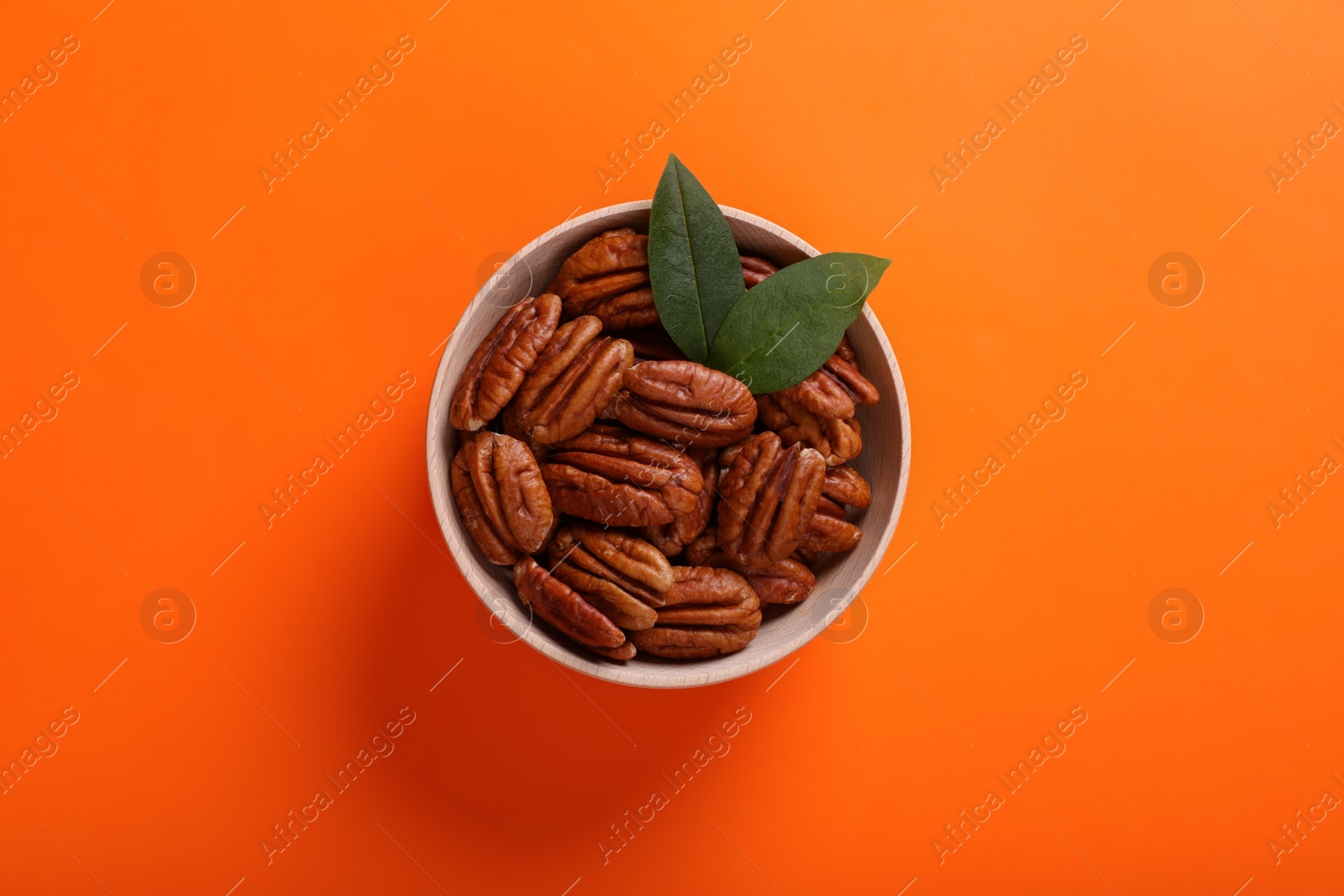 Photo of Bowl with tasty pecan nuts and green leaves on orange background, top view