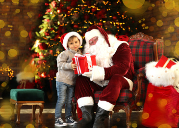 Santa Claus giving present to little boy near Christmas tree indoors