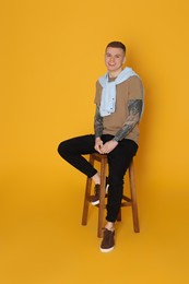 Photo of Smiling young man with tattoos sitting on stool against yellow background