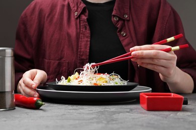 Photo of Stir-fry. Woman with chopsticks eating tasty rice noodles with meat and vegetables at grey textured table, closeup