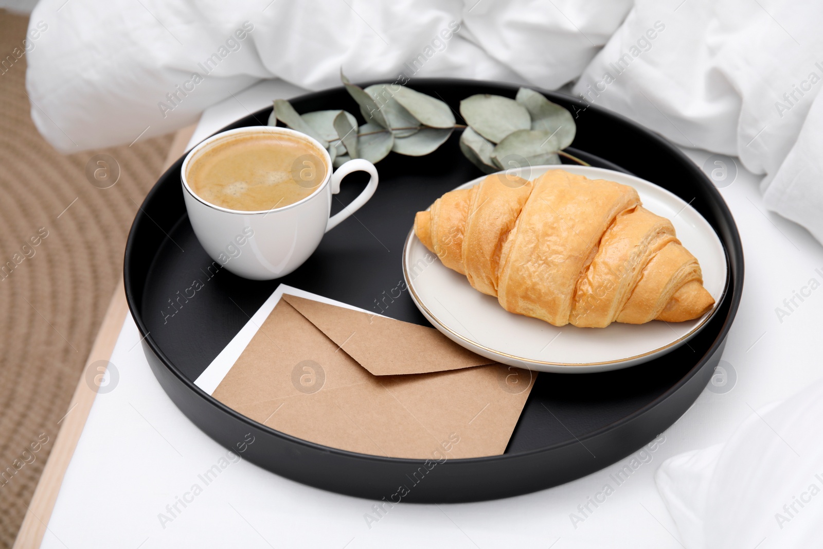 Photo of Tray with tasty croissant, cup of coffee and envelope on white bed