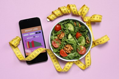Smartphone with weight loss calculator application, measuring tape and bowl of salad on pink background, flat lay