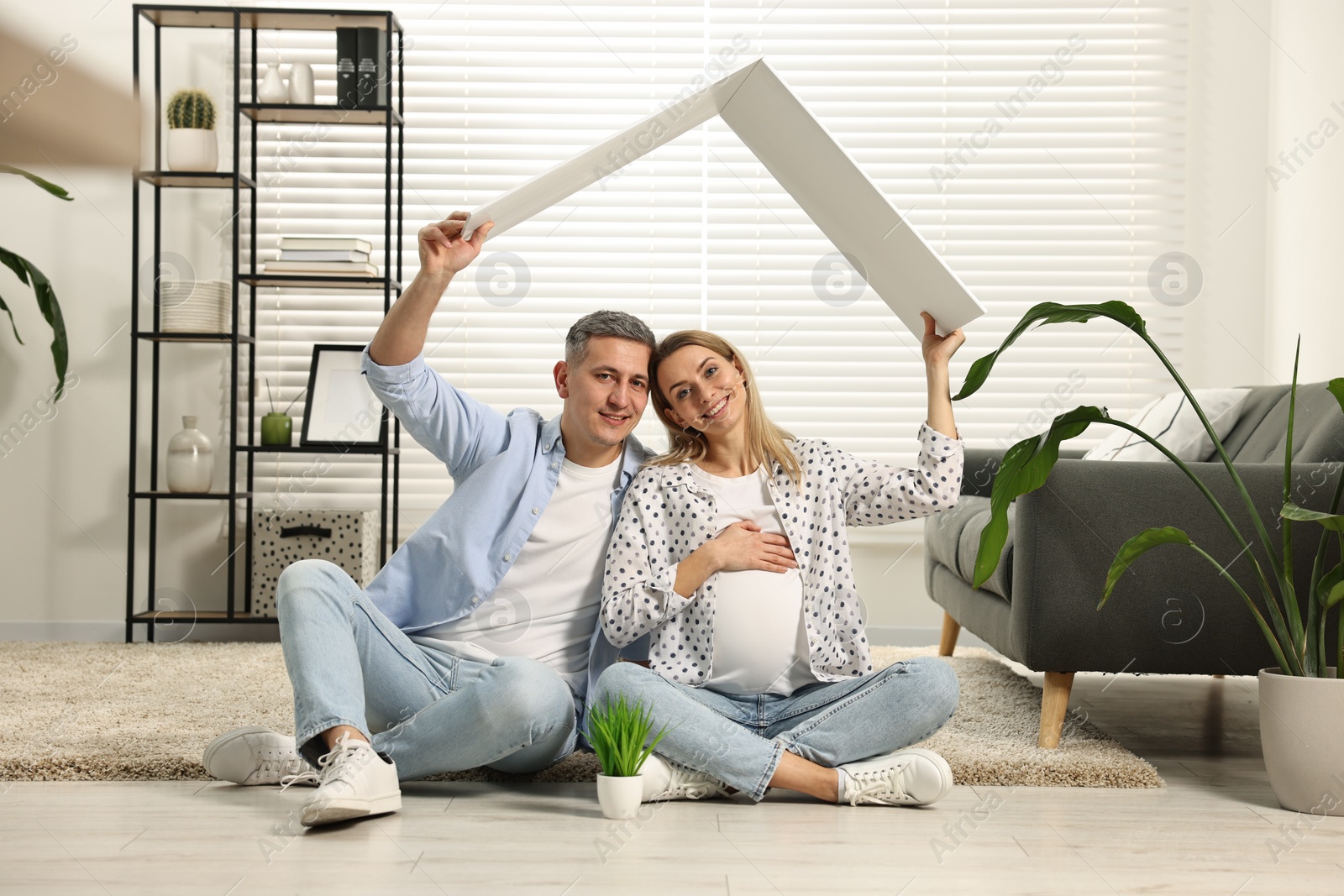 Photo of Young family housing concept. Pregnant woman with her husband sitting under cardboard roof on floor at home